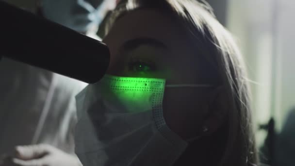 Une femme portant un masque médical protecteur vérifie ses yeux lors d'un rendez-vous chez l'ophtalmologiste. Un laser vert brille dans la pupille. Traitement ophtalmologique - une jeune femme vérifie son acuité visuelle — Video