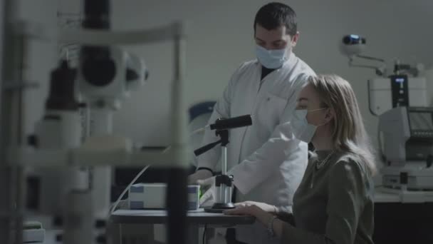 Uma mulher usando uma máscara médica protetora verifica os olhos em uma consulta oftalmológica. Um laser verde brilha na pupila. Tratamento oftalmológico - uma jovem mulher verificando sua acuidade visual — Vídeo de Stock