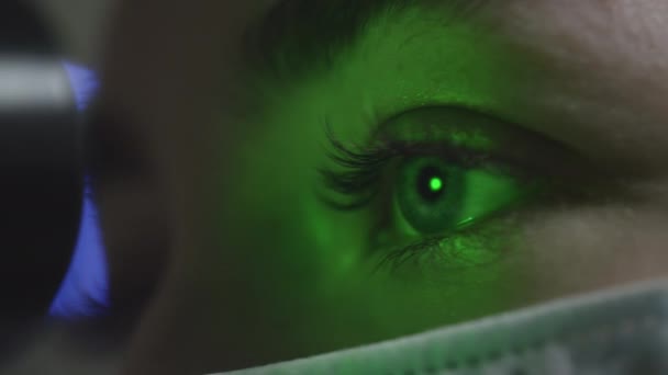 Una mujer que lleva una máscara médica se revisa los ojos en una cita con un oftalmólogo. Un láser verde brilla en la pupila. Tratamiento oftalmológico: una mujer joven que comprueba su agudeza visual — Vídeo de stock