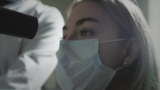 Een vrouw met een beschermend medisch masker controleert haar ogen bij een oogheelkundige afspraak. Een groene laser schijnt in de pupil. oogheelkundige behandeling - een jonge vrouw die haar gezichtsscherpte controleert — Stockvideo