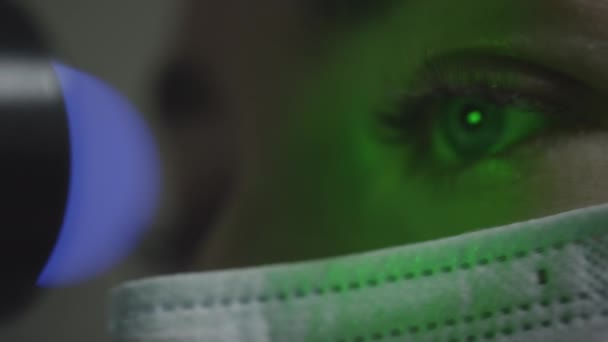 Een vrouw met een medisch masker controleert haar ogen bij een oogheelkundige afspraak. Een groene laser schijnt in de pupil. oogheelkundige behandeling - een jonge vrouw die haar gezichtsscherpte controleert — Stockvideo