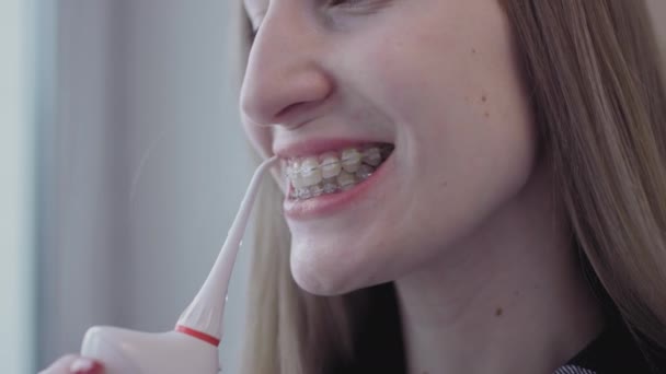 Mujer limpia aparatos ortopédicos con riego dental con hilo de agua. La chica se encarga del sistema de frenos en sus dientes. Limpia los dientes con hilo de agua portátil dientes dentales eléctricos limpiador dental riego oral — Vídeos de Stock