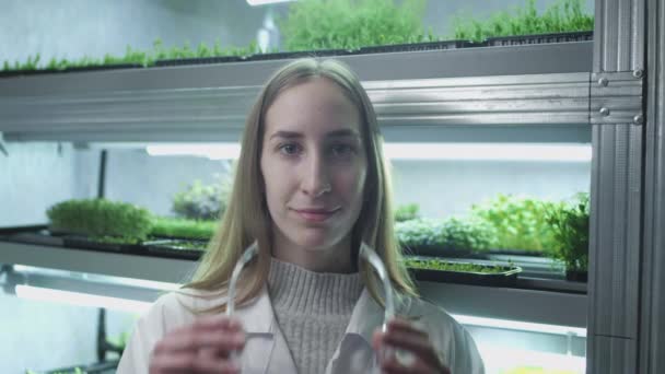 Wissenschaftlerin im weißen Kittel in einem Botaniklabor. Das Mädchen setzt sich eine Plastikbrille auf. Studentinnen züchten experimentelle gentechnisch veränderte Pflanzen. Konzept: Forschung, Biochemie — Stockvideo