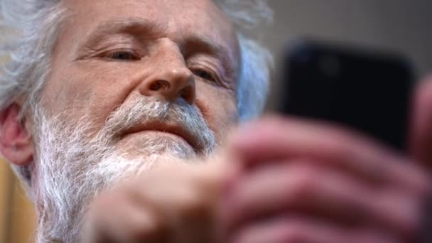 Сіро-волохатий старий з бородою тримає в руці смартфон, використовує його. Дід дивиться на фотографії в гаджеті. Зробити відеодзвінок або замовити їжу онлайн вдома. Написання блогу про здоров'я . — стокове відео