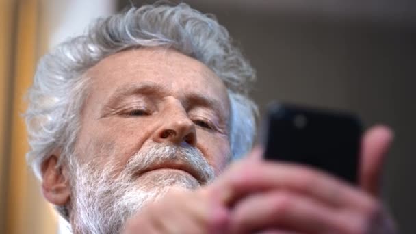 Un anciano canoso con barba sostiene un smartphone en la mano, lo usa. El abuelo mira fotos en un gadget. Haz una videollamada u ordena comida en línea en casa. Escribe un blog sobre salud. — Vídeos de Stock