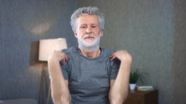 Omuzlarında egzersiz yapan gri saçlı son sınıf öğrencisinin portresi. Arka planda yapılan ev sporları hareketleri. Büyükbabam mükemmel bir atletik vücut şekline sahip. Spor giyimli yaşlı adam..