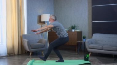 Tek bacağında sakalı olan gri saçlı yaşlı bir adamın portresi. Yoga minderinin üstünde duruyor. Ev sporları egzersizleri. Büyükbabam mükemmel bir atletik vücut şekline sahip. Spor giyimli yaşlı adam..