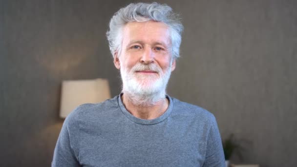 Un anciano caucásico de pelo gris mira a la cámara, sonríe. Retrato del viejo feliz europeo en casa. Edad de oro abuelo cuidado de la salud estilo de vida alegre, libertad relajar el concepto de jubilación. — Vídeo de stock