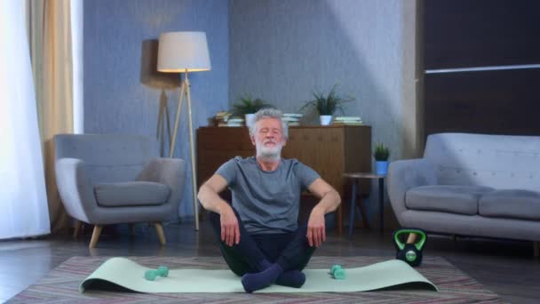 El retrato de un hombre mayor de pelo gris con barba se dedica al yoga en la esterilla. Se sienta en la posición de loto, asana. Ojos cerrados, inhala profundamente. Abuelo en excelente forma corporal atlética. — Vídeos de Stock