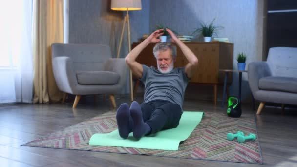 A gray-haired senior man with a beard does a press exercise, pumps up the abdominal muscles. In the living room, doing exercise on a yoga mat. Old man in sportswear — Stock Video