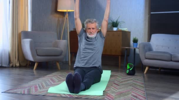 Un bătrân cu părul gri, cu barbă, stă pe un covor de yoga. Bunicul din sufragerie face un exercițiu cu gantere, ridică mâinile în sus. Bătrân în îmbrăcăminte sportivă. Ai grijă la corpul tău la bătrânețe . — Videoclip de stoc