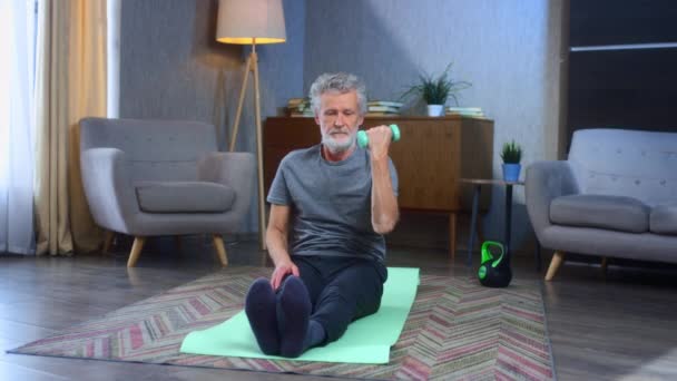 Portrait of a gray-haired senior man with a beard goes in for sports at home with dumbbells. In the living room exercises for pumping up the muscles of the arm beats. Old man in sportswear — Stock Video