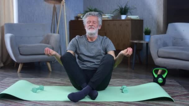 Portretul unui bătrân cu părul gri, cu barbă, este implicat în yoga pe covor. Stă în poziţia de lotus, asana. Ochii închişi, inhalează adânc. Bunicul în formă excelentă de corp atletic . — Videoclip de stoc