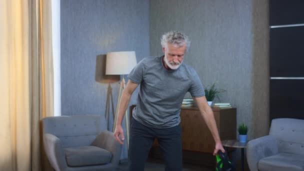 Homme âgé aux cheveux gris avec une barbe fait de l'exercice de fitness avec une kettlebell. Le grand-père dans le salon soulève un poids. Vieil homme en tenue de sport. Succès et réussite après un travail acharné. — Video