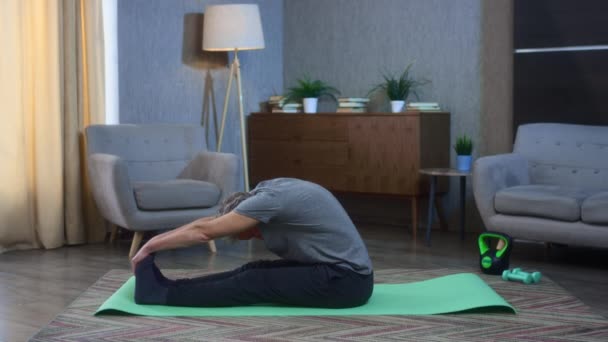 Hombre mayor de pelo gris se estira en casa en una esterilla de yoga. Ejercicio casero para los músculos de la espalda y las piernas. El abuelo extiende los brazos hasta los pies. Viejo en ropa deportiva. Salud en la vejez — Vídeos de Stock