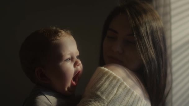 The child is in her mothers arms, yawns widely and looks with blue eyes. Sleepy baby. Millennial mother holds her son in her arms by the window of the house. A childs face in the sun. — Stock Video