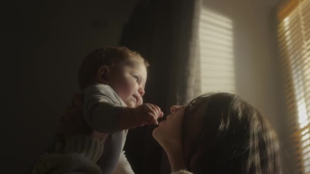 Een kind in de armen van een lachend meisje. Millennial moeder kotst in haar zoons armen voor het raam van het huis. Blinds op het raam. De kinderen kijken in de zon. Warme gele toon. Gezinswaarden. — Stockvideo