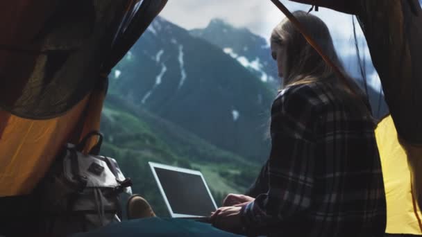 Uma jovem freelancer usa um laptop para trabalhar ao ar livre. Trabalho remoto em uma tenda amarela perto das grandes montanhas. — Vídeo de Stock