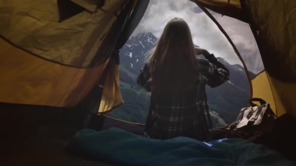 一个穿着格子呢衬衫的女徒步旅行者坐在一个黄色的帐篷里休息。看那些大山. — 图库视频影像