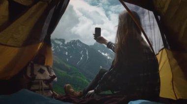 Ekose gömlek giymiş bir yürüyüşçü kız sarı çadırda oturur, akıllı telefondan selfie çeker.