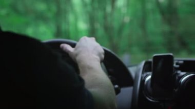 Yeşil bir ormanda araba süren bir adam. Kırışık ellerle siyah deri bir direksiyona yakın çekim.