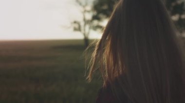 Kırmızı tişörtlü genç bir kız gün batımında ya da şafakta bir tarlada duruyor. Saçlar rüzgârla savruluyor.