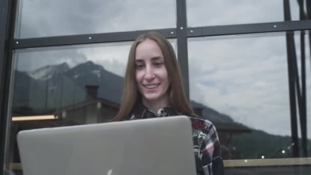 Ένα νεαρό όμορφο κορίτσι χρησιμοποιεί ένα φορητό υπολογιστή για μια βιντεοκλήση σε ένα καφέ με φόντο τα βουνά. Επαγγελματική κλήση ή βίντεο συνάντηση με τους γονείς — Αρχείο Βίντεο