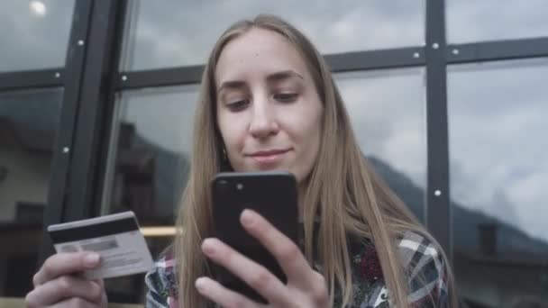 Een jong mooi meisje gebruikt een smartphone en een bankkaart in een café tegen de achtergrond van besneeuwde bergen — Stockvideo