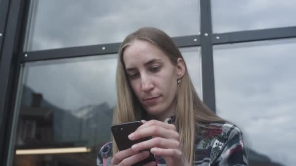 Ung vacker flicka använder en smartphone i ett café på bakgrunden av snötäckta berg. Nedflyttning under karantän och pandemi — Stockvideo