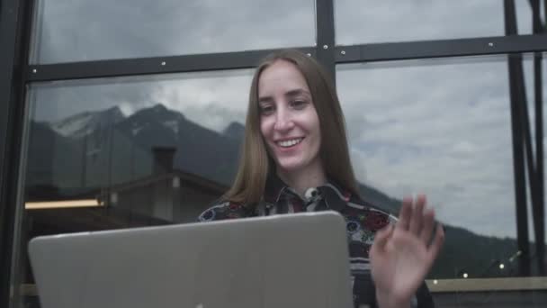 En ung vacker flicka använder en bärbar dator för ett videosamtal i ett café mot bakgrund av bergen. Affärssamtal eller videomöte med föräldrar — Stockvideo