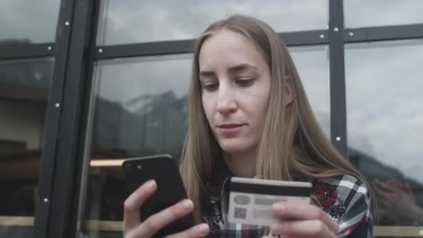 Junge schöne Mädchen benutzt Smartphone und Bankkarte in einem Café vor der Kulisse der Berge — Stockvideo