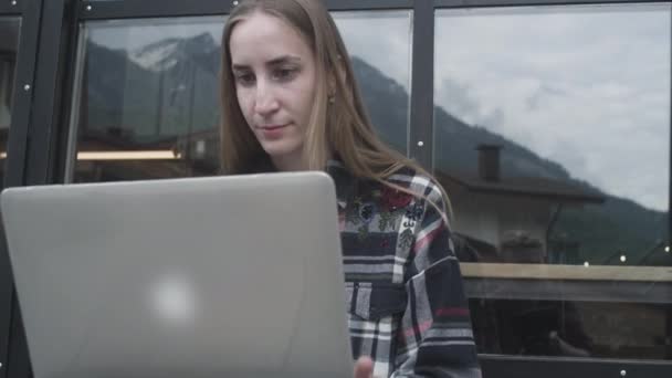 En ung frilansarflicka jobbar på en laptop på ett café nära bergen. Kvinnan skriver på en bärbar dator, program. — Stockvideo