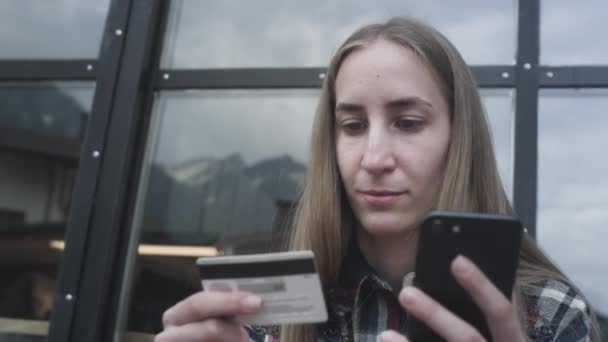 En ung vacker flicka använder en smartphone och ett bankkort i ett café mot bakgrund av snötäckta berg — Stockvideo