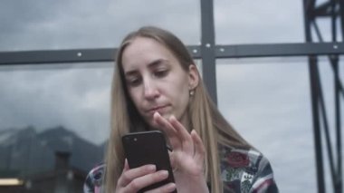 Cep telefonuyla çitte oturan gülümseyen kadın. Yüksek dağların turistik yolu. Dijital iletişim. Sosyal ağdaki arkadaşlarla mesajlaşma