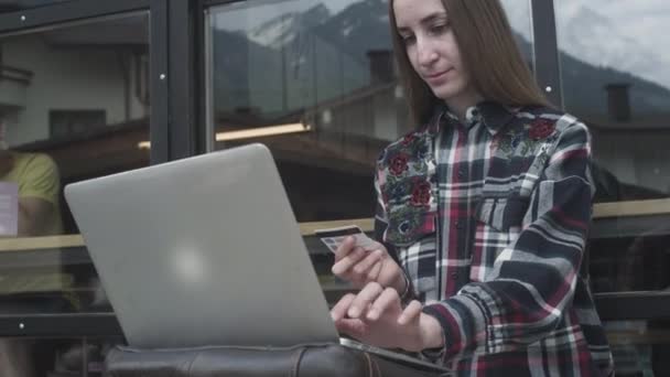 En ung vacker flicka använder en bärbar dator och ett bankkort i ett café mot bakgrund av bergen — Stockvideo