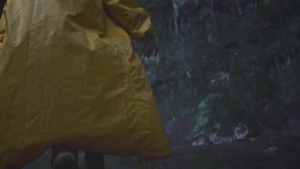 Ung flicka resenär vandrare i en gul regnrock promenader till ett vattenfall i höglandet, höjer händerna. Njuta av naturen i bergen, resa. — Stockvideo