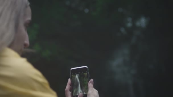 Caucásico Chica viajero en un impermeable amarillo hace una foto de una cascada en un teléfono inteligente en el bosque. — Vídeos de Stock