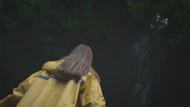 Ung flicka resenär vandrare i en gul regnrock promenader till ett vattenfall i höglandet, höjer händerna. Njuta av naturen i bergen, resa. — Stockvideo
