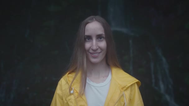 Portret van een blanke meisjesreiziger in een gele regenjas bij een waterval in het bos. Lifestyle concept, in slow motion — Stockvideo