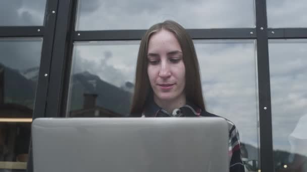 Μια νεαρή ελεύθερη επαγγελματίας δουλεύει σε ένα λάπτοπ σε ένα καφέ κοντά στα βουνά. Η γυναίκα πληκτρολογεί σε ένα φορητό υπολογιστή, προγράμματα. — Αρχείο Βίντεο