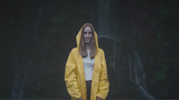 Retrato de viajante Caucasiano Menina em uma capa de chuva amarela perto de uma cachoeira na floresta. Conceito de estilo de vida, em câmera lenta — Vídeo de Stock