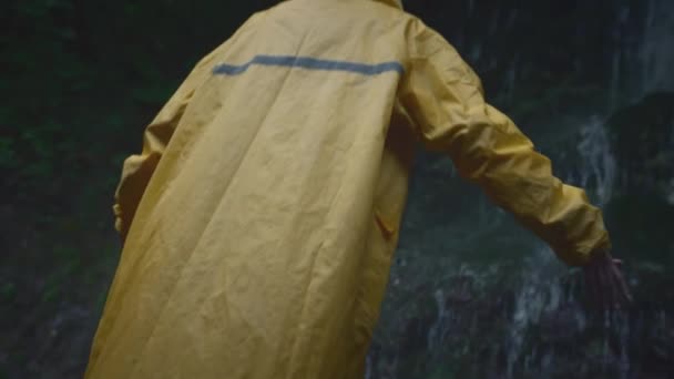 Ein junges Wandermädchen im gelben Regenmantel geht zu einem Wasserfall im Hochland, hebt die Hände, genießt die Natur und das Leben. Reisen in den Bergen, Abenteuer auf Reisen. Lifestyle-Konzept. — Stockvideo