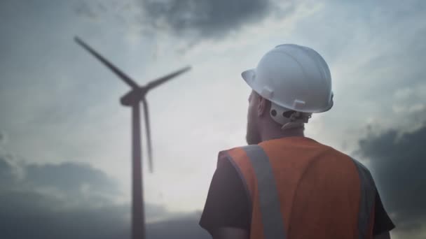 PORTRÄT: Ein männlicher Ingenieur oder Arbeiter mit Helm und Weste neben einer Windkraftanlage. Ein junger Mann mit Bart, weißem Schutzhelm und Weste — Stockvideo