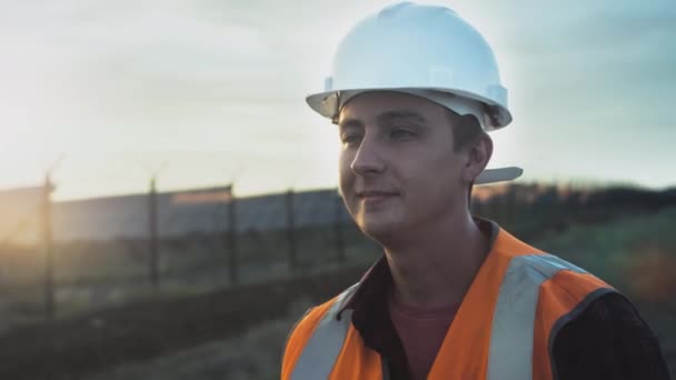 Gün batımında güneş panellerinin yanında genç bir mühendisin portresi. Yenilenebilir enerji kaynaklarının sağlığını ve verimliliğini kontrol eder. — Stok video