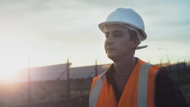 Gün batımında güneş panellerinin yanında genç bir mühendisin portresi. Yenilenebilir enerji kaynakları ve güneş pillerinin sağlığını ve verimliliğini kontrol eder. Güneş paneli plantasyonu. Yeşil enerji kavramı — Stok video