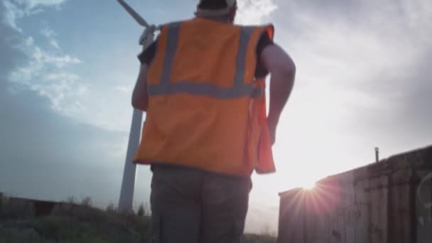 Een man met een witte helm en een vest rent naar een windturbine die een ongeluk repareert. Ingenieur of werknemer. Inspecteert en past de schroef aan — Stockvideo