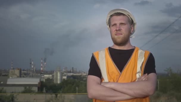 RETRATO: Um engenheiro ou trabalhador do sexo masculino em um capacete e um colete na frente de uma fábrica ou produção. Um antigo manufactory industrial polui o ar. — Vídeo de Stock