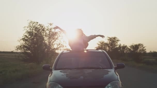 Νεαρό όμορφο κορίτσι βόλτες στο αυτοκίνητο με τα χέρια ψηλά, μια γυναίκα στην ηλιοροφή του αυτοκινήτου. Απολαμβάνοντας τη ζωή στο ηλιοβασίλεμα σε μια καταπακτή αυτοκινήτου — Αρχείο Βίντεο