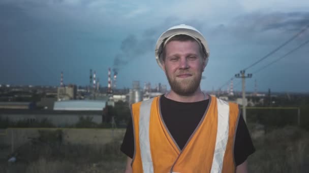 PORTRAIT: Egy férfi mérnök vagy munkás sisakban és mellényben egy üzem vagy gyár előtt Egy régi ipari manufaktúra szennyezi a levegőt — Stock videók