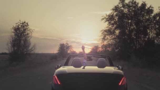 Νεαρό κορίτσι οδηγεί ένα κάμπριο, σηκώνει το χέρι της πάνω κατά τη διάρκεια του ηλιοβασιλέματος, χειρονομώντας. Κουνώντας το χέρι κατά την οδήγηση. Γυναίκα οδηγεί αυτοκίνητο. — Αρχείο Βίντεο